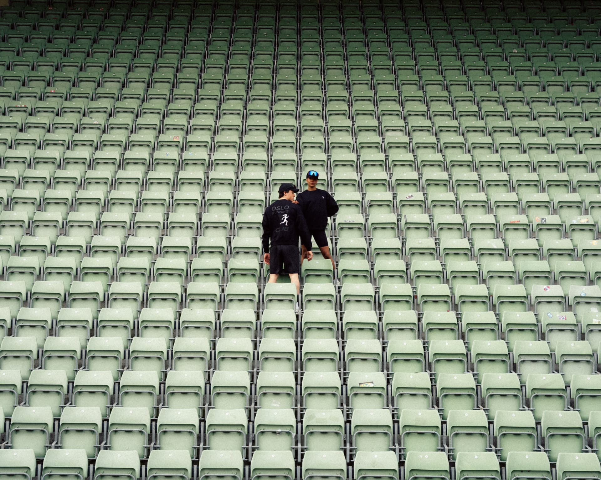Two people in black activewear standing among rows of pale green stadium seats